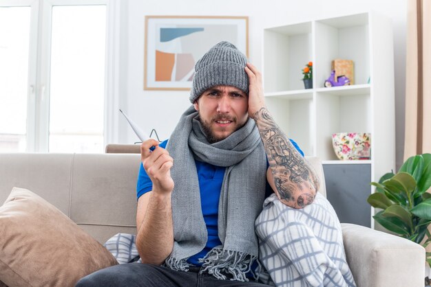 Aching young ill man wearing scarf and winter hat sitting on sofa in living room holding thermometer  keeping hand on head