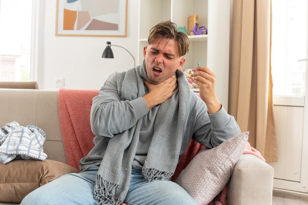 aching young ill man with scarf around neck putting hand on his neck and holding thermometer sitting on couch at living room