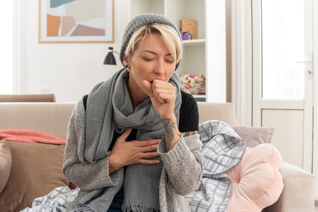 Aching young ill slavic woman with scarf around her neck wearing winter hat coughing keeping fist close to mouth sitting on couch at living room