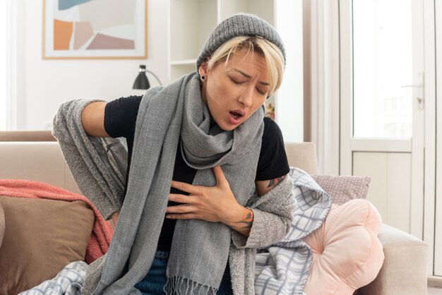 Aching young ill slavic woman with scarf around her neck wearing winter hat putting hand on her chest and on her back sitting on couch at living room