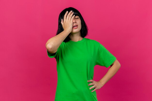 Aching young ill woman putting hand on face keeping hand on waist with closed eyes isolated on pink wall with copy space