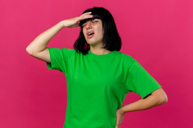 Aching young ill woman putting hand on head and on waist with closed eyes isolated on pink wall