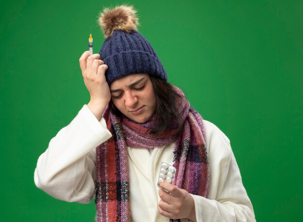 ローブの冬の帽子と注射器と錠剤のパックを身に着けている痛む若い病気の女性は、緑の壁に隔離された目を閉じて頭に手を保ちます