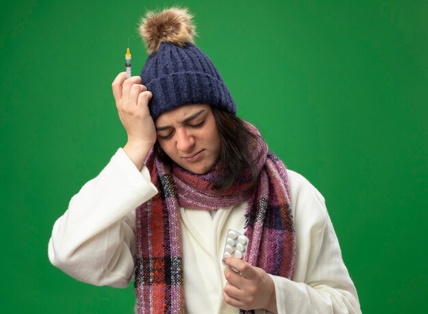 Aching young ill woman wearing robe winter hat and scarf holding syringe and pack of tablets keeping hand on head with closed eyes isolated on green wall