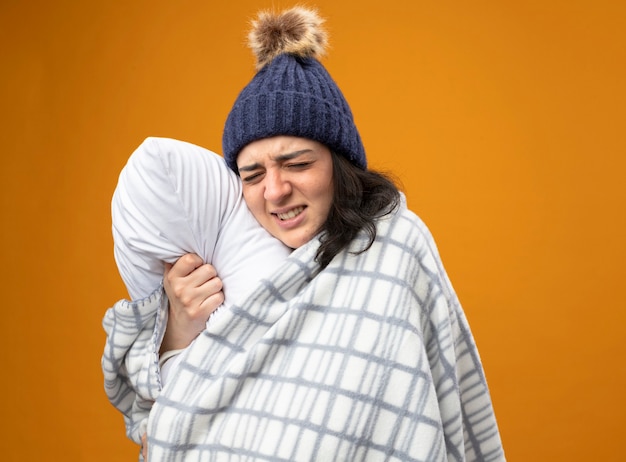 Aching young ill woman wearing robe winter hat wrapped in plaid standing in profile view hugging pillow with closed eyes isolated on orange wall