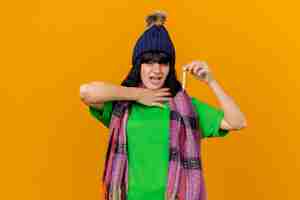無料写真 冬の帽子とスカーフを身に着けて、オレンジ色の壁に隔離された喉に手を置いて温度計を見て痛む若い病気の女性