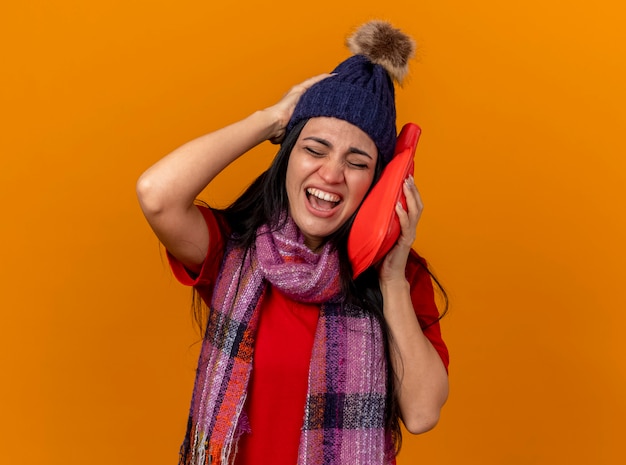Болит молодая больная женщина в зимней шапке и шарфе, касающаяся лица мешком с горячей водой, держащая руку за голову с закрытыми глазами, изолированными на оранжевой стене