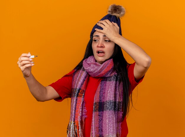 Aching young ill woman wearing winter hat and scarf holding and looking at thermometer putting hand on head isolated on orange wall