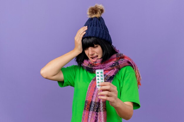 Aching young ill woman wearing winter hat and scarf holding pack of capsules keeping hand on head suffering from headache with closed eyes isolated on purple wall with copy space