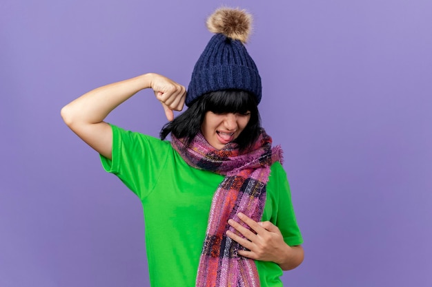 Foto gratuita giovane donna malata dolorante che indossa cappello invernale e sciarpa tenendo la mano sullo stomaco e il pugno in aria con gli occhi chiusi isolati sulla parete viola con lo spazio della copia