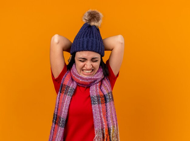 Aching young ill woman wearing winter hat and scarf keeping hands behind neck with closed eyes isolated on orange wall
