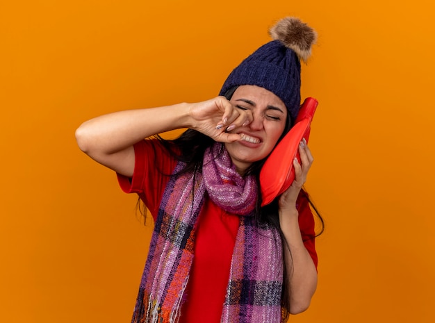 Aching young ill woman wearing winter hat and scarf touching face with hot water bag touching nose with closed eyes isolated on orange wall