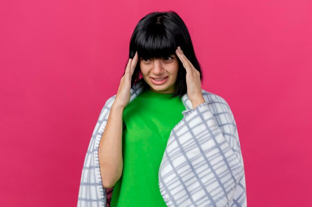 Aching young ill woman wrapped in plaid keeping hands on head looking straight isolated on pink wall with copy space