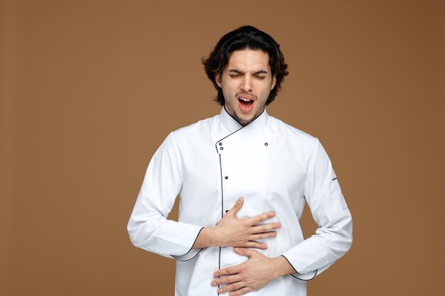 aching young male chef wearing uniform keeping hands on belly screaming with closed eyes isolated on brown background