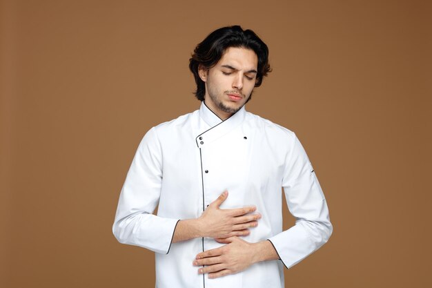 aching young male chef wearing uniform keeping hands on belly with closed eyes isolated on brown background