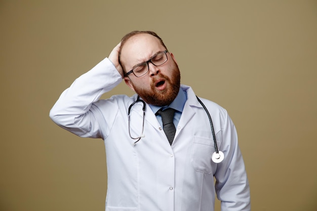 Aching young male doctor wearing glasses lab coat and stethoscope around his neck keeping hand on head with closed eyes isolated on olive green background