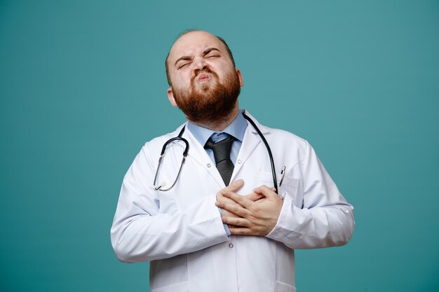 Aching young male doctor wearing medical coat and stethoscope around his neck keeping hands on heart having heartache with closed eyes isolated on blue background