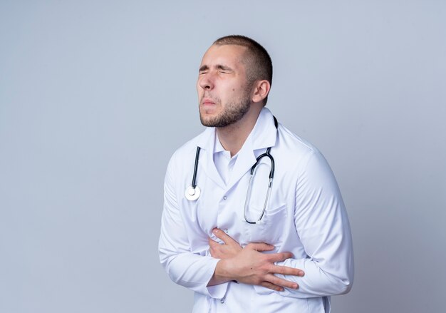 Aching young male doctor wearing medical robe and stethoscope around his neck holding his belly suffering from pain with closed eyes isolated on white  with copy space