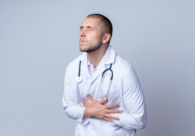 Aching young male doctor wearing medical robe and stethoscope around his neck holding his belly with closed eyes isolated on white  with copy space