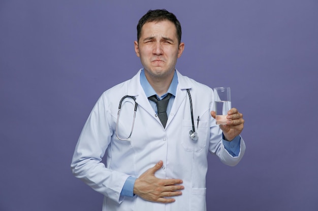 Aching young male doctor wearing medical robe and stethoscope around neck holding glass of water looking at camera while keeping hand on belly isolated on purple background