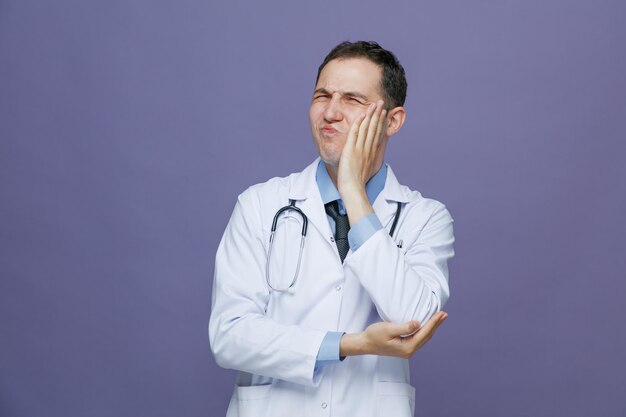 aching young male doctor wearing medical robe and stethoscope around neck keeping hand under elbow and another hand on face with closed eyes having toothache isolated on purple background
