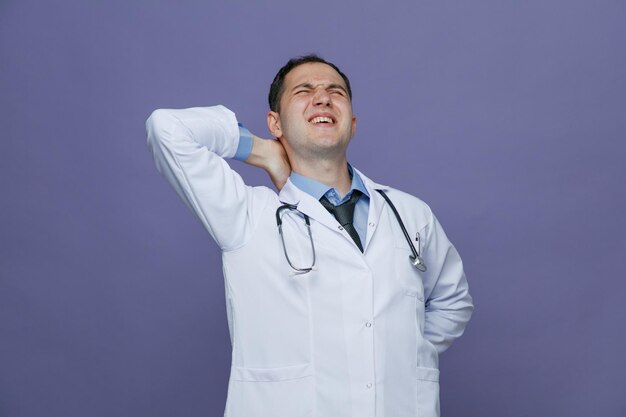 Больной молодой врач-мужчина в медицинском халате и стетоскопе на шее держит руки на спине и за шеей с закрытыми глазами, изолированными на фиолетовом фоне