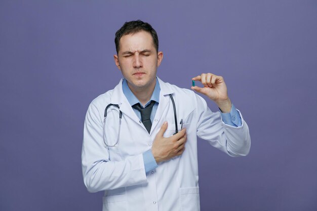 Aching young male doctor wearing medical robe and stethoscope around neck showing capsule with closed eyes while keeping hand on heart isolated on purple background