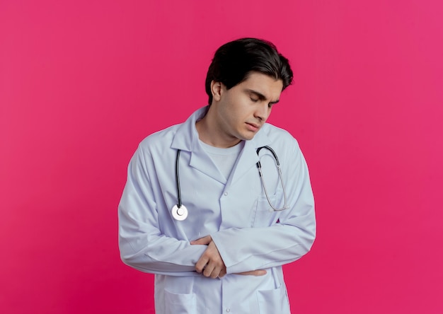 Aching young male doctor wearing medical robe and stethoscope putting hand on belly and holding wrist with closed eyes isolated on pink wall with copy space