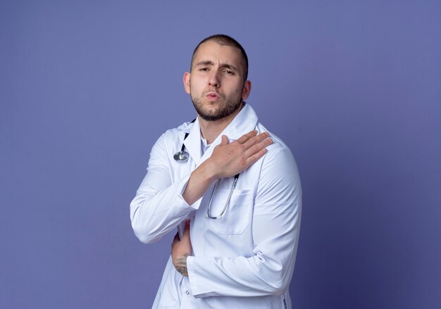 Aching young male doctor wearing medical robe and stethoscope putting hand on shoulder and on kidney isolated on purple  with copy space