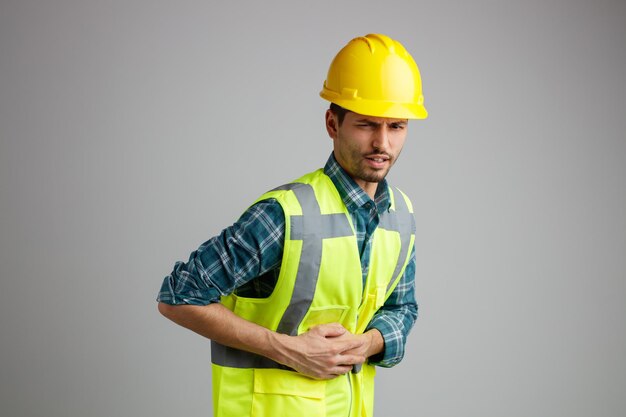 Aching young male engineer wearing safety helmet and uniform standing in profile view looking at camera keeping hands on belly isolated on white background with copy space