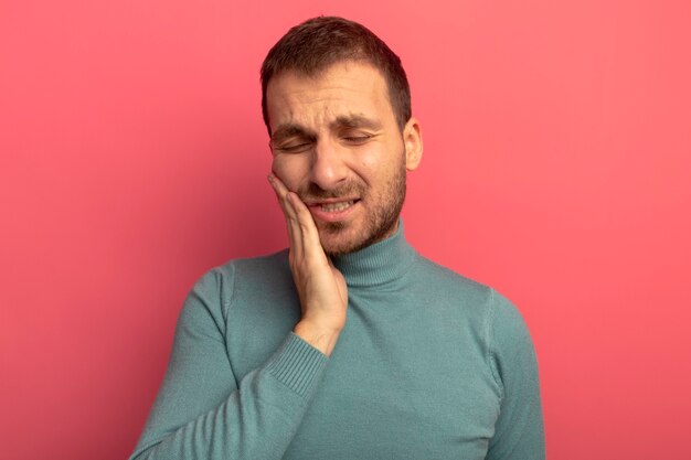 Aching young man putting hand on cheek suffering from headache with closed eyes isolated on pink wall