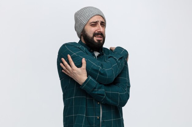 Aching young man wearing winter hat looking at camera keeping hands on arm and on shoulder isolated on white background