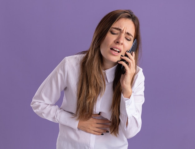 Aching young pretty caucasian girl puts hand on belly and talks on phone isolated on purple wall with copy space