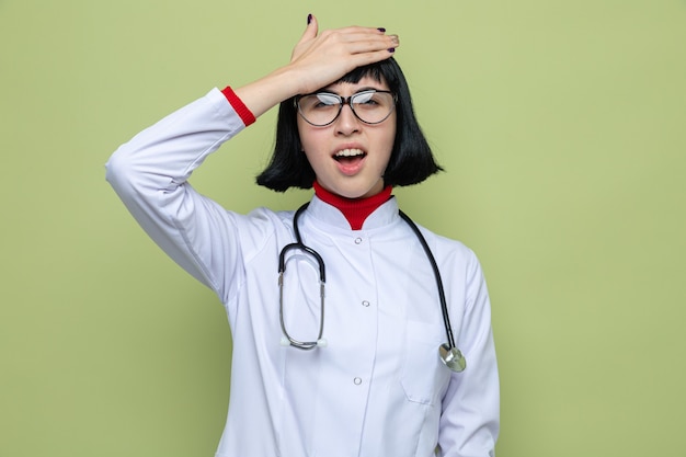 Una giovane ragazza caucasica dolorante con occhiali ottici in uniforme da medico con stetoscopio mette la mano sulla testa e guarda la telecamera