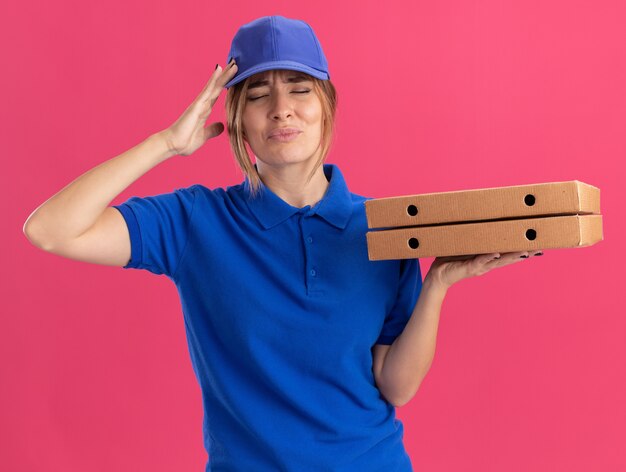 Aching young pretty delivery woman in uniform puts hand on head and holds pizza boxes isolated on pink wall