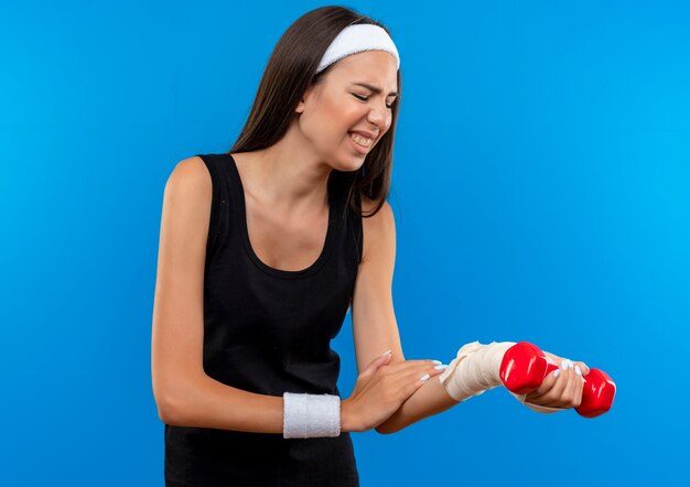 Aching young pretty sporty girl wearing headband and wristband holding dumbbell putting hand on and looking at her injured wrist wrapped with bandage isolated on blue wall
