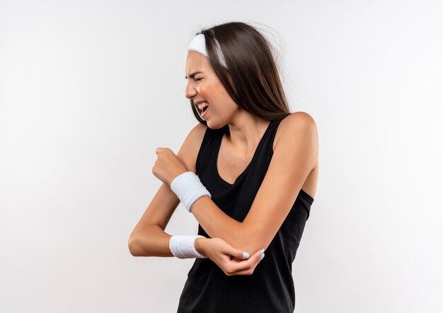 Aching young pretty sporty girl wearing headband and wristband putting hand on arm with closed eyes on white wall with copy space