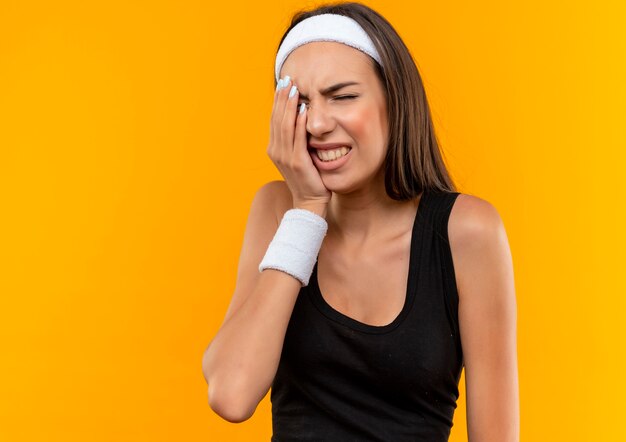 Aching young pretty sporty girl wearing headband and wristband putting hand on face with closed eyes isolated on orange wall with copy space