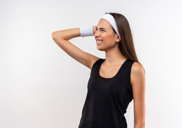 Aching young pretty sporty girl wearing headband and wristband putting hand on head looking at side on white wall with copy space