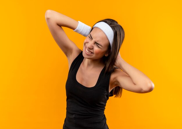 Aching young pretty sporty girl wearing headband and wristband putting hands behind her head on orange wall