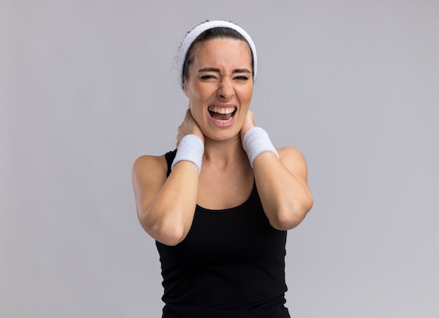 Aching young pretty sporty woman wearing headband and wristbands keeping hands on neck