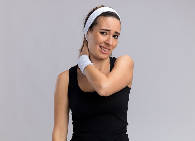 Aching young pretty sporty woman wearing headband and wristbands looking at front putting hand on neck isolated on white wall with copy space