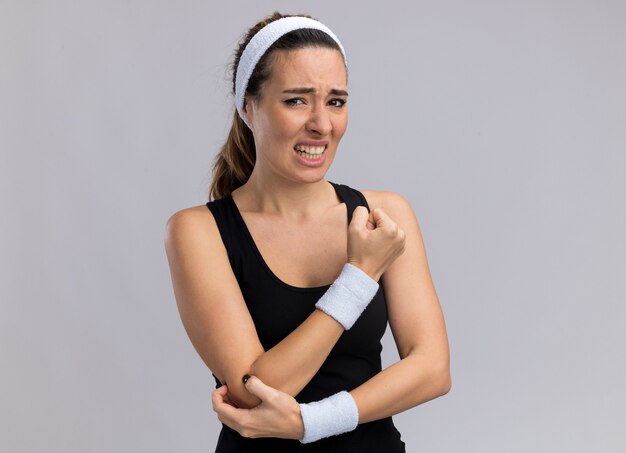 Aching young pretty sporty woman wearing headband and wristbands touching elbow