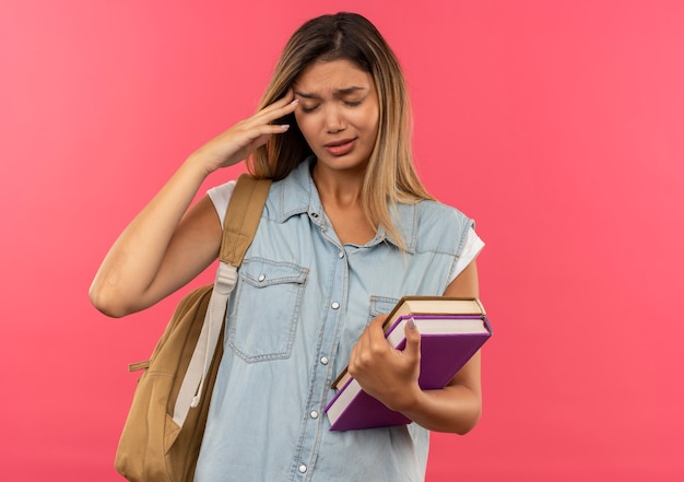 Aching young pretty student girl wearing back bag holding books and putting hand on temple suffering from headache with closed eyes isolated on pink  with copy space