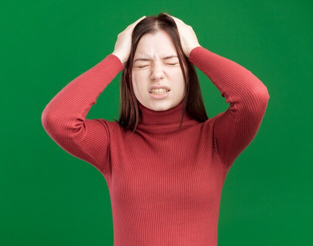 Aching young pretty woman keeping hands on head with closed eyes isolated on green wall