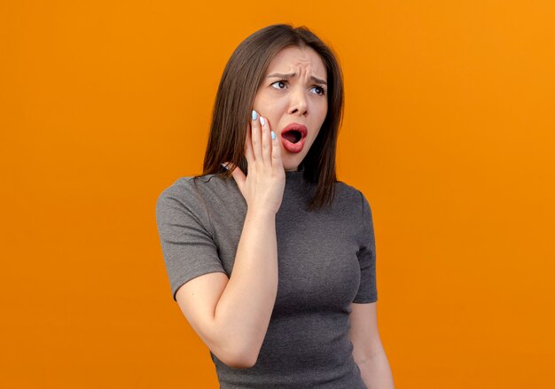 Aching young pretty woman looking straight and putting hand on cheek suffering from toothache isolated on orange background with copy space