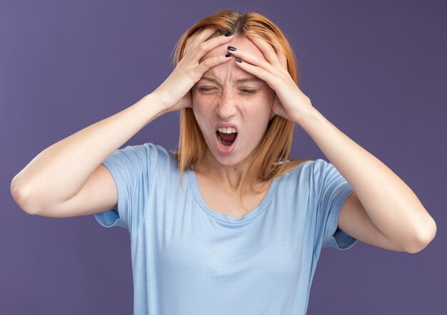 Aching young redhead ginger girl with freckles puts hands on forehead on purple