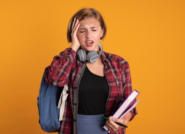 Free photo aching young slavic student girl with headphones wearing backpack puts hand on head holds book and notebook