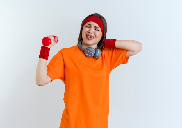 Aching young sporty woman wearing headband and wristbands and headphones on neck holding dumbbell putting hand behind neck with closed eyes isolated