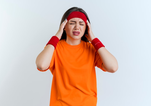 Aching young sporty woman wearing headband and wristbands holding head suffering from headache with closed eyes isolated on white wall with copy space